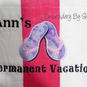 May include: A beach towel with pink and white stripes. The towel features an embroidered design of two flip-flops with a tie-dye pattern in shades of purple and blue. The text "Ann's Permanent Vacation" is embroidered in black.