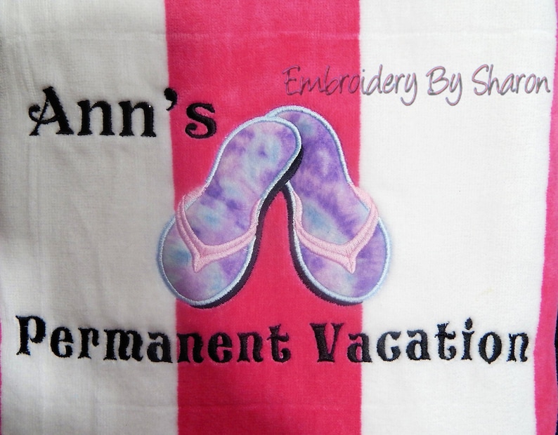 May include: A beach towel with pink and white stripes. The towel features an embroidered design of two flip-flops with a tie-dye pattern in shades of purple and blue. The text "Ann's Permanent Vacation" is embroidered in black.