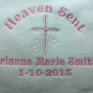 May include: White blanket with pink embroidered text and a cross design. The text reads "Heaven Sent" arched across the top, with the name "Arianna Marie Smith" and the date "1-10-2013" below the cross. The cross is detailed with a pink center and silver outline.