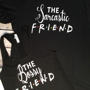 K&ouml;nnte beinhalten: Zwei schwarze Oberteile mit wei&szlig;em Text. Eines ist ein T-Shirt mit der Aufschrift "The Sarcastic FRIEND", das andere ist ein Tanktop mit der Aufschrift "The Bossy FRIEND". Das Wort "FRIEND" ist im Stil der Fernsehsendung Friends.