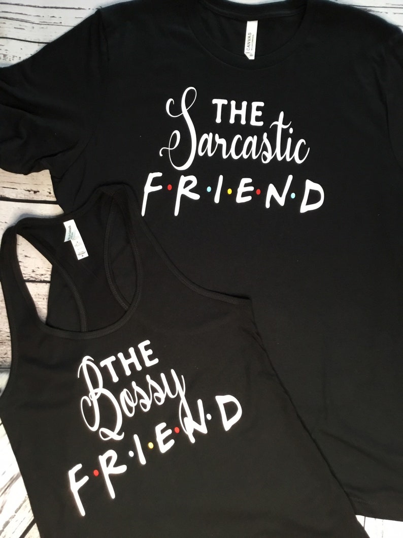 K&ouml;nnte beinhalten: Zwei schwarze Oberteile mit wei&szlig;em Text. Eines ist ein T-Shirt mit der Aufschrift "The Sarcastic FRIEND", das andere ist ein Tanktop mit der Aufschrift "The Bossy FRIEND". Das Wort "FRIEND" ist im Stil der Fernsehsendung Friends.