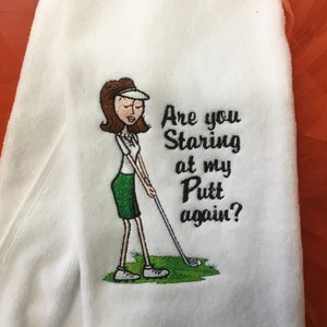 May include: White golf towel with an embroidered cartoon of a woman golfer. The woman is wearing a visor, white shirt, and green skirt, and is holding a golf club. Text reads "Are you Staring at my Putt again?"