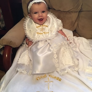 Peut inclure: Un b&eacute;b&eacute; portant une robe de bapt&ecirc;me en satin blanc et un bavoir assorti, orn&eacute;s de dentelle. Le bavoir et la couverture sont brod&eacute;s avec "God Bless Vivian Grace November 18, 2018". Le b&eacute;b&eacute; porte un bandeau blanc.