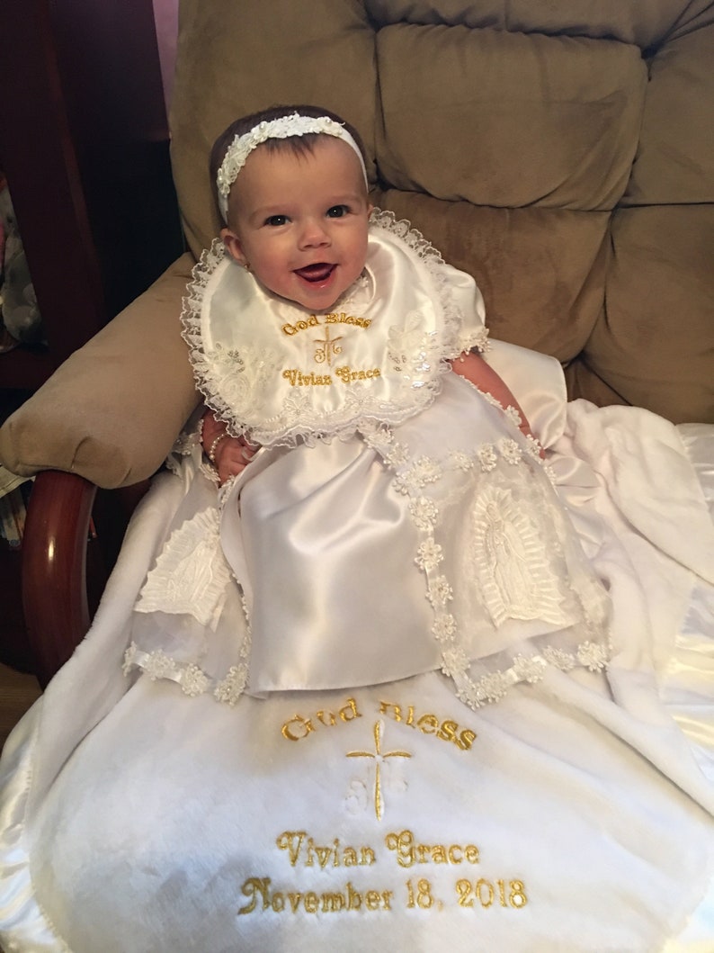 Peut inclure: Un b&eacute;b&eacute; portant une robe de bapt&ecirc;me en satin blanc et un bavoir assorti, orn&eacute;s de dentelle. Le bavoir et la couverture sont brod&eacute;s avec "God Bless Vivian Grace November 18, 2018". Le b&eacute;b&eacute; porte un bandeau blanc.