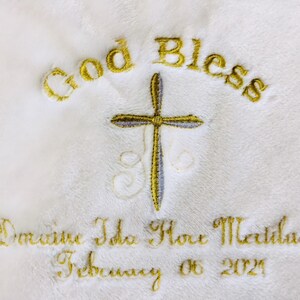 May include: White embroidered blanket with gold text reading "God Bless" above a cross. Below the cross, the blanket is embroidered with a name and the date "February 06 2021". The embroidery is in gold thread.
