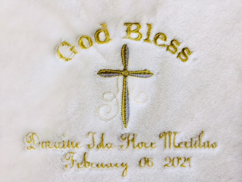 May include: White embroidered blanket with gold text reading "God Bless" above a cross. Below the cross, the blanket is embroidered with a name and the date "February 06 2021". The embroidery is in gold thread.
