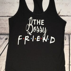 K&ouml;nnte beinhalten: Schwarzes Tanktop mit Racerback-Design und dem wei&szlig;en Schriftzug "THE Bossy FRIEND" in Schreib- und Blockschrift. Das Wort "FRIEND" hat bunte Punkte anstelle der Buchstaben "I" und "E". Das Tanktop ist aus weichem Material.
