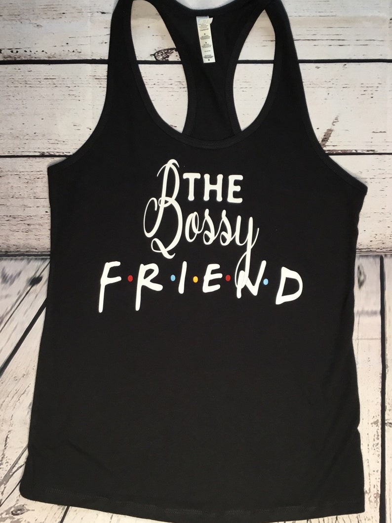 K&ouml;nnte beinhalten: Schwarzes Tanktop mit Racerback-Design und dem wei&szlig;en Schriftzug "THE Bossy FRIEND" in Schreib- und Blockschrift. Das Wort "FRIEND" hat bunte Punkte anstelle der Buchstaben "I" und "E". Das Tanktop ist aus weichem Material.