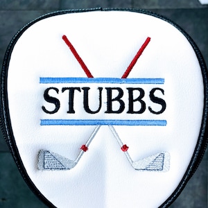 May include: White golf club head cover with the name "STUBBS" embroidered in black, with blue and red accents. Two crossed golf clubs are embroidered below the name. The cover has a black border.