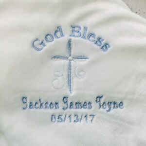 May include: White baby blanket with blue embroidered text that reads "God Bless", a cross, and "Jackson James Toyne 05/13/17". The blanket has a soft, plush texture.