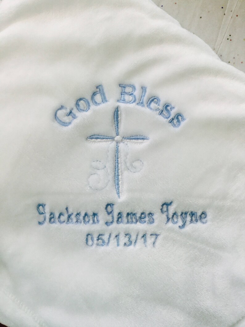 May include: White baby blanket with blue embroidered text that reads "God Bless", a cross, and "Jackson James Toyne 05/13/17". The blanket has a soft, plush texture.