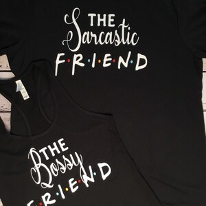 K&ouml;nnte beinhalten: Schwarzes T-Shirt und Tanktop mit wei&szlig;em Text. Das T-Shirt liest "THE Sarcastic FRIEND" und das Tanktop "THE Bossy FRIEND". Das Wort "FRIEND" ist im Stil der Fernsehsendung Friends mit farbigen Punkten.