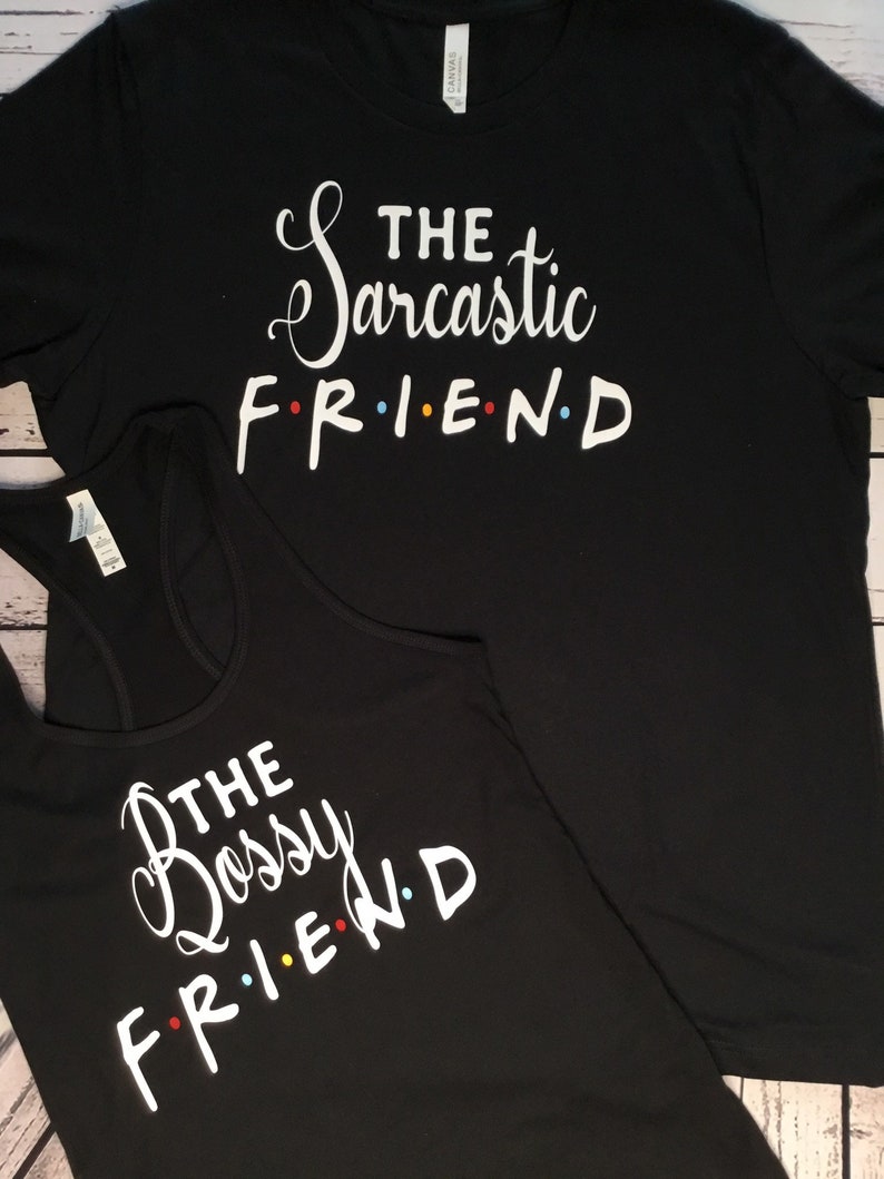 K&ouml;nnte beinhalten: Schwarzes T-Shirt und Tanktop mit wei&szlig;em Text. Das T-Shirt liest "THE Sarcastic FRIEND" und das Tanktop "THE Bossy FRIEND". Das Wort "FRIEND" ist im Stil der Fernsehsendung Friends mit farbigen Punkten.