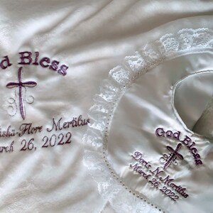 May include: White baby blanket and bib set. The blanket and bib are embroidered with the words "God Bless", a cross, and the name "Doris Nishi-Flore Mertilus" and the date "March 26, 2022". The bib has a lace trim.