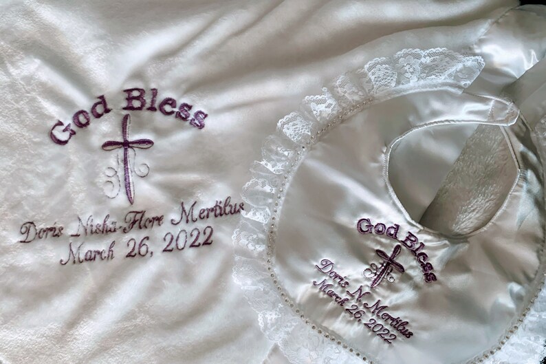 May include: White baby blanket and bib set. The blanket and bib are embroidered with the words "God Bless", a cross, and the name "Doris Nishi-Flore Mertilus" and the date "March 26, 2022". The bib has a lace trim.