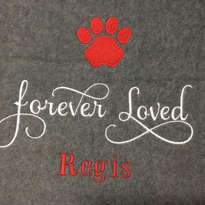 May include: A gray fleece blanket with a red paw print and the embroidered text "forever Loved" in white script. Below, the name "Regis" is embroidered in red. A comforting pet memorial.
