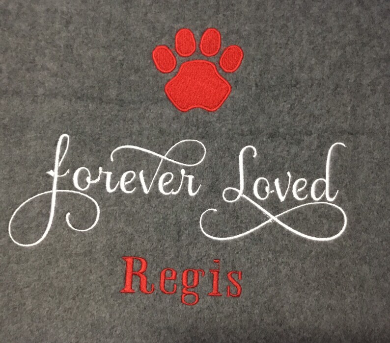 May include: A gray fleece blanket with a red paw print and the embroidered text "forever Loved" in white script. Below, the name "Regis" is embroidered in red. A comforting pet memorial.