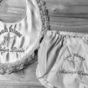 May include: A white satin baby bib and matching bloomers set. Both items are embroidered with the words "God Bless" above a cross and the name "Madelyn Lizzie". The bib has a scalloped lace trim.