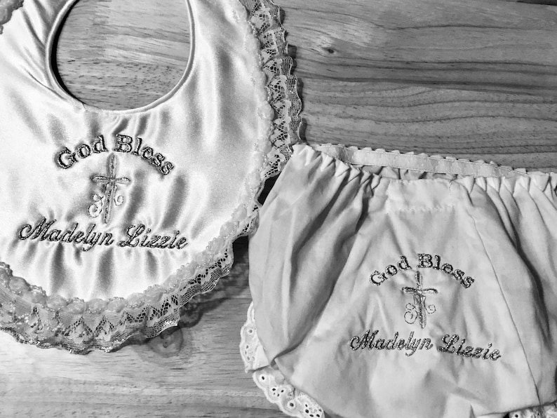 May include: A white satin baby bib and matching bloomers set. Both items are embroidered with the words "God Bless" above a cross and the name "Madelyn Lizzie". The bib has a scalloped lace trim.
