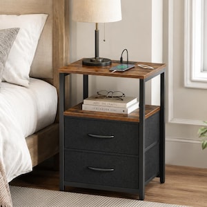 May include: A square nightstand with a dark brown wood-grain top and shelf, featuring two black fabric drawers with curved handles. The frame is made of black metal, and the top surface includes an integrated power outlet with a charging cable.