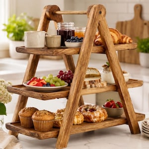 May include: A three-tiered wooden serving stand with an A-frame design. The shelves hold various food items including muffins, pastries, fresh fruit, and small bowls. The structure is made of natural wood with a visible grain and a sturdy handle at the top.