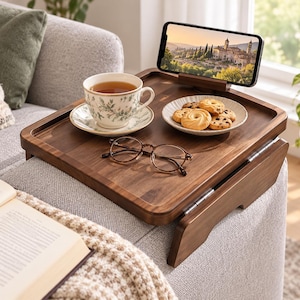 May include: A rectangular wooden sofa arm tray with a dark brown finish, featuring a built-in smartphone stand, a ceramic teacup and saucer, a small plate of cookies, and a pair of brown-rimmed eyeglasses resting on the flat surface.