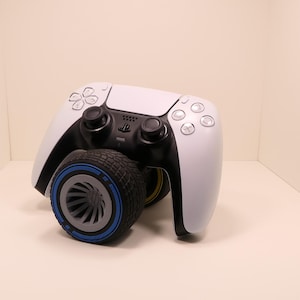 May include: A white and black video game controller featuring a circular black tire-shaped attachment on the front. The tire has a grey center with a blue ring accent. The controller includes standard buttons, two analog sticks, and a central logo.