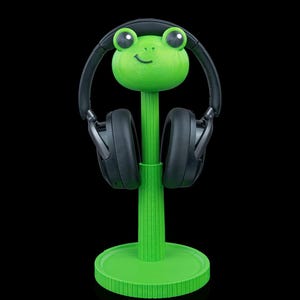 May include: A bright green frog-shaped headphone stand with a circular base and a vertical post. The frog head features large eyes and a small smile. A pair of black over-ear headphones is resting on the stand, supported by the frog's head.