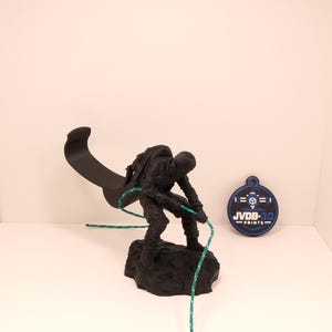 Pode incluir: Uma estatueta impressa em 3D preto fosco de um escalador sobre uma base rochosa, com uma extensão de gancho curva e uma corda verde. Ao lado da estatueta há uma etiqueta circular azul e preta com o texto branco JVDB-3D PRINTS e um ícone.
