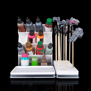 May include: A white tiered paint organizer holding various plastic bottles of acrylic paint, alongside a white rectangular base with multiple vertical wooden sticks featuring metal clips that hold small gray and purple miniature figures.