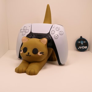 Op de afbeelding: Een bruine controllerstandaard in de vorm van een kat die een witte en zwarte gamecontroller vasthoudt. Het katfiguur heeft grote zwarte ogen, een kleine mond en lichte binnenoren. Een klein rond zwart label met de witte tekst JVDB is zichtbaar.