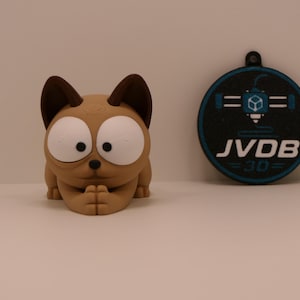 Op de afbeelding: Een beige kattenfiguurtje met grote witte ogen en donkerbruine oren, naast een rond zwart en blauw medaillon met een 3D-printerpictogram en de tekst JVDB 30. De kat heeft de voorpoten bij elkaar in een rusthouding.