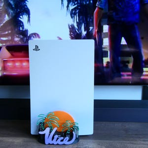 Pode incluir: Uma consola PlayStation 5 branca com um acessório decorativo na base. O acessório apresenta um sol laranja brilhante, palmeiras verdes e a palavra Vice num tipo de letra cursivo roxo, sobre uma silhueta preta de paisagem urbana.