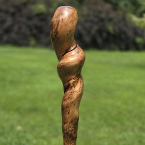 May include: A tall, slender wooden walking stick featuring a polished, light brown finish with visible natural wood grain. The top section is carved into a distinct, spiraling, twisted shape that tapers to a smooth, rounded point at the very top.