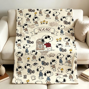 May include: A cream-colored fleece blanket featuring a repeating pattern of Snoopy wearing sunglasses and a Joe Cool shirt, interspersed with small yellow birds, a jukebox, and a central banner with the text NAME in a serif font.