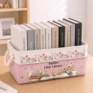 Peut inclure: Un panier de rangement rectangulaire en tissu rose avec des poignées en corde blanche. Le panier présente une bordure florale sur le haut et un motif imprimé d'un livre ouvert, de fleurs et d'une tasse avec le texte "Sophia's Little Library".