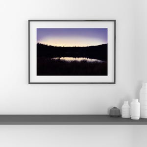 May include: A framed landscape photograph featuring a dark silhouette of a forest and tall grasses bordering a calm lake at twilight. The sky transitions from a deep purple at the top to a soft, glowing yellow horizon line above the tree line.