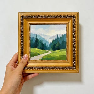 May include: A square oil painting in an ornate gold frame depicting a landscape with a winding dirt path leading through a vibrant green meadow toward a dense forest of dark pine trees under a soft, cloudy sky. The text ARTSPAINTSBYWAJAHAT is visible.