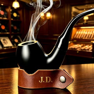 May include: A smooth black smoking pipe with a curved stem, emitting wisps of white smoke. The pipe rests in a brown leather holder featuring the embossed initials J.D. and a single silver-toned metal snap button closure on the side.