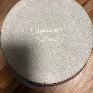 May include: The circular base of a ceramic vessel featuring a speckled light grey surface. The text Edgecomb Potters is inscribed in white cursive lettering across the centre of the base, which has a smooth, matte texture and a dark brown rim.