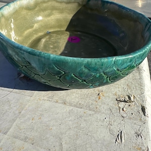 May include: A round ceramic bowl featuring a textured exterior with overlapping scale-like patterns in shades of deep teal and green. The interior of the bowl has a smooth, light olive-green glaze and contains a small, circular purple sticker.