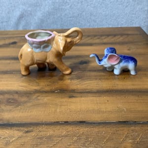 May include: A tan ceramic elephant figurine with a pink and light blue saddle-shaped planter on its back, positioned next to a smaller blue and white ceramic elephant figurine featuring pink ears and a trunk raised in an upward position.