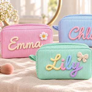 May include: Three rectangular zippered pouches with striped fabric. The pink pouch features the name Emma and a white flower. The blue pouch displays the name Chloe and a pink heart. The green pouch shows the name Lily and a beige bow.