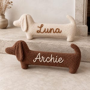 May include: Two plush dachshund-shaped pillows made of textured fabric. The top pillow is cream-colored with the name Luna embroidered in brown. The bottom pillow is brown with the name Archie embroidered in cream-colored thread.