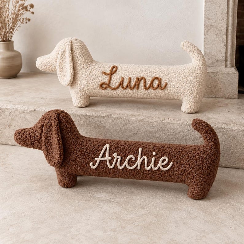 May include: Two plush dachshund-shaped pillows made of textured fabric. The top pillow is cream-colored with the name Luna embroidered in brown. The bottom pillow is brown with the name Archie embroidered in cream-colored thread.