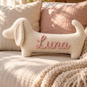May include: A cream-colored, textured, dog-shaped throw pillow featuring the name Luna written in pink yarn script on its side. The pillow has a soft, nubby surface, a long body, floppy ears, and an upright tail, crafted in a plush material.