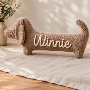 May include: A brown, textured, sherpa-style plush toy shaped like a dachshund dog. The side of the plush features the name Winnie written in white, raised, cursive lettering. The toy has floppy ears, a long body, and an upright tail.
