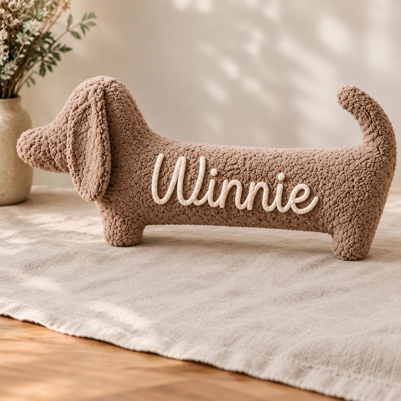 May include: A brown, textured, sherpa-style plush toy shaped like a dachshund dog. The side of the plush features the name Winnie written in white, raised, cursive lettering. The toy has floppy ears, a long body, and an upright tail.