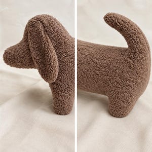 May include: A brown plush dog toy made of textured, curly fabric. The toy features a long body, a rounded head with a long, floppy ear, and a curved tail. The design is segmented into two parts, showing the front section and the rear section.