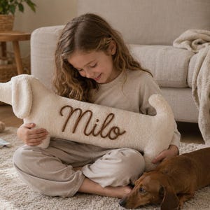 May include: A cream-colored, plush, dachshund-shaped pillow featuring the name Milo embroidered in brown script lettering on its side. The pillow has a soft, textured fabric surface, a long body, floppy ears, and a small tail.